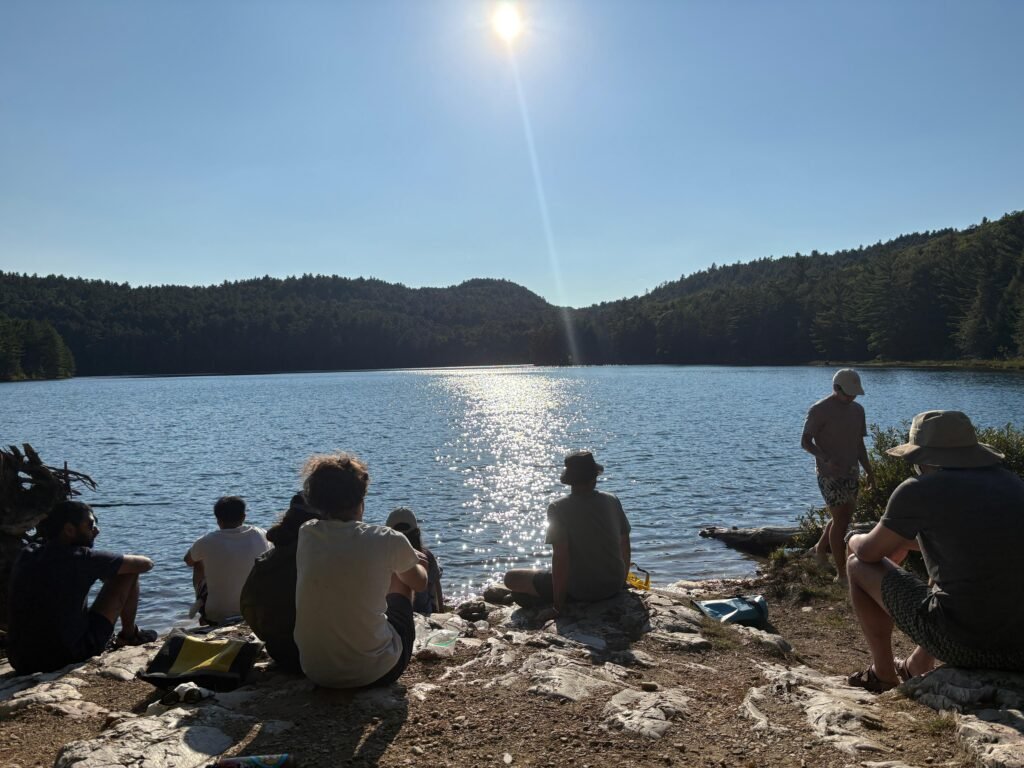 Group of people sitting on a rocky shoreline in Killarney watching the sun reflect across a calm lake surrounded by forested hills. The scene captures a quiet moment at camp with gear nearby as they relax by the water at the end of the day.