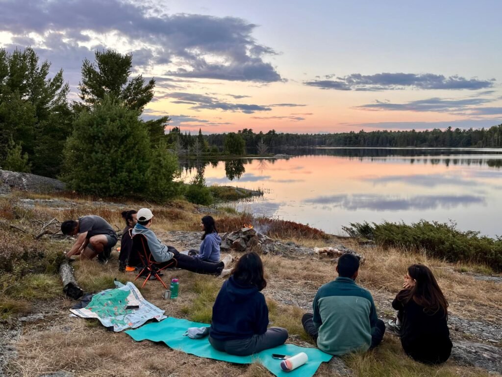 Group of friends sitting around a small campfire on a rocky lakeside campsite in Killarney with calm water reflecting a pastel sunset sky. Camping mats, chairs, and gear are spread out as they relax and take in the evening view.