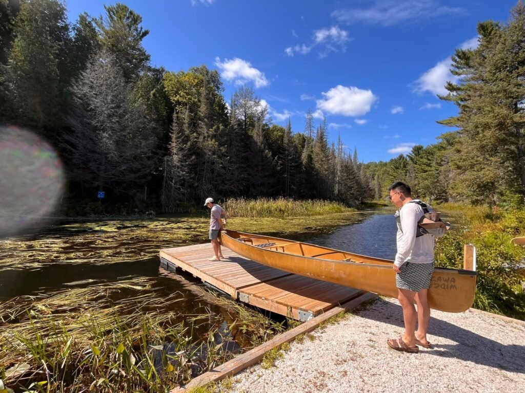 Two people carrying a canoe onto a small wooden dock at a narrow waterway in Killarney surrounded by dense forest and lily pads. The scene shows a typical portage or launch moment as they prepare to continue their canoe route.