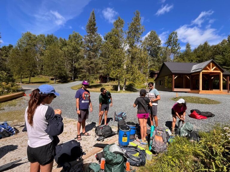 Group of friends organizing backpacks and canoe camping gear in a gravel parking area surrounded by forest at a Killarney access point. Packs, barrels, and paddles are spread out as they prepare for a backcountry canoe trip under a bright blue sky. Displays ultralight backpacking gear.