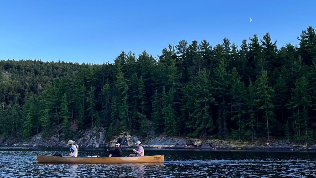 Three people paddling a canoe across a calm lake in Killarney with a dense pine forest and rocky shoreline in the background under a clear evening sky. The still water and soft light capture the peaceful backcountry experience of canoe camping in the park.