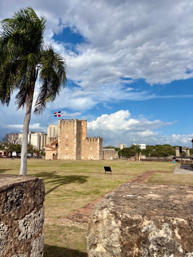 Historic stone fortress with the Dominican Republic flag flying above it and a tall palm tree beside the walls under dramatic clouds in Santo Domingo. The open grassy grounds and colonial architecture highlight one of the historic landmarks visitors see when exploring things to do in Santo Domingo.