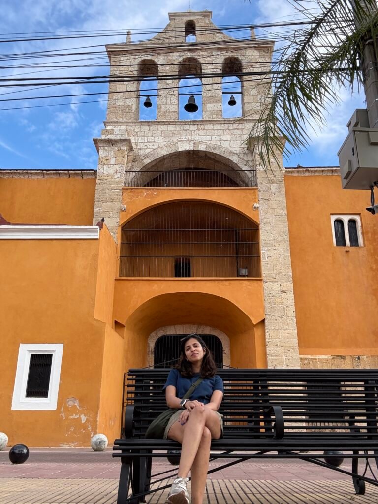 Woman sitting on a black bench in front of a bright orange colonial church with a stone bell tower and three hanging bells in Santo Domingo. The historic architecture in the Zona Colonial is one of the cultural highlights people visit when looking for things to do in Santo Domingo.