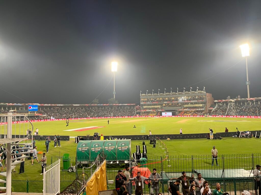 Gaddafi Stadium in Lahore with its large circular structure and bright floodlights towering over the cricket field. Crowds gather in the stands reflecting the city’s deep passion for cricket and shared experiences.