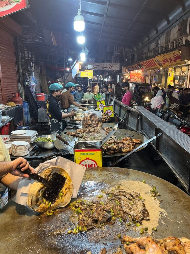 A large metal tawa at Arif Chatkhara sizzles with pieces of chicken being cooked in spices by a street food vendor. Flames and smoke rise around the pan highlighting Lahore’s rich and lively food scene.