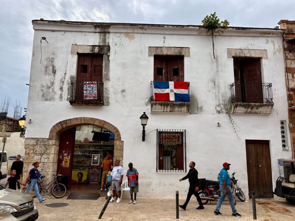 Weathered white colonial building in Santo Domingo with dark wooden balconies and a Dominican Republic flag hanging above a busy street. Locals walk past small storefronts and parked motorcycles beneath the historic facade in the Colonial Zone.