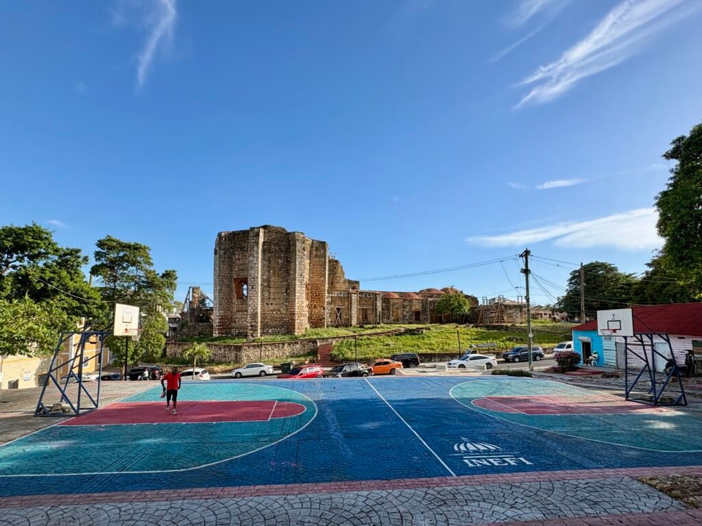 Outdoor basketball courts in Santo Domingo with bright blue and red pavement set in front of historic stone ruins under a clear blue sky. A few people stand near the courts while cars and trees surround the open plaza.
