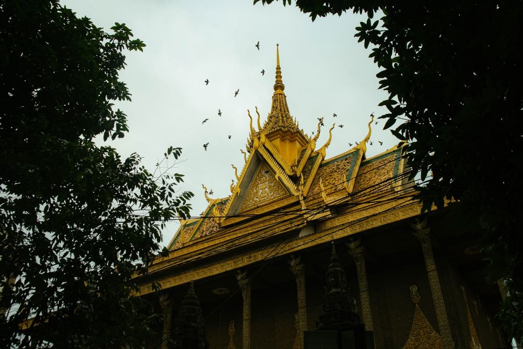 A majestic golden pagoda in Phnom Penh with birds flying above at sunset, surrounded by lush trees.