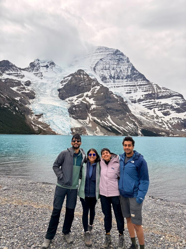Four hikers stand on the rocky shoreline of Berg Lake with turquoise water, the Berg Glacier, and snow-covered Mount Robson towering behind them.