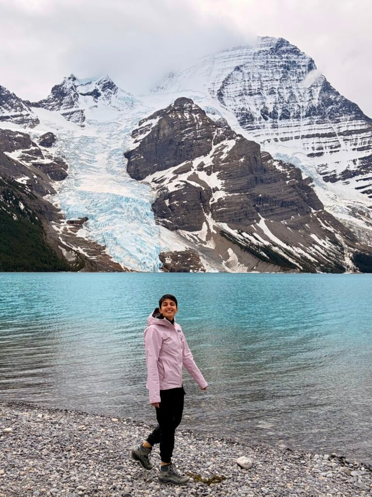 Hiker standing on the rocky shore of Berg Lake with turquoise water stretching toward the Berg Glacier and snow-covered peaks of Mount Robson. The glacier descends directly toward the lake, showcasing the spectacular scenery that awaits at the end of the Berg Lake Trail.
