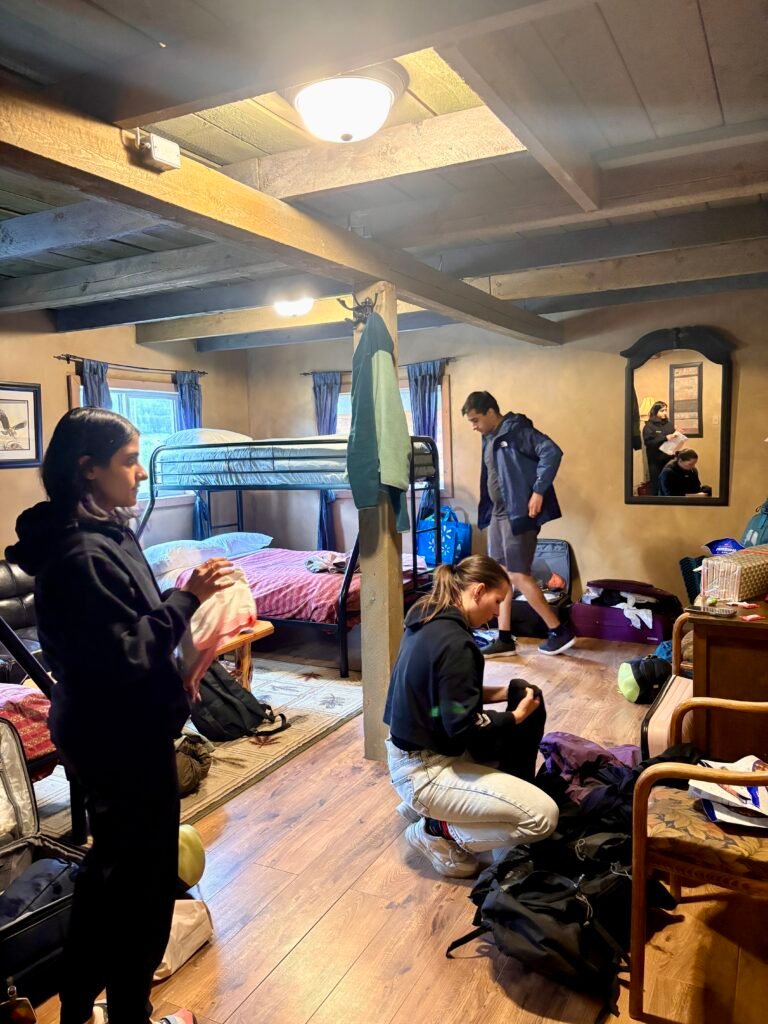 Rustic cabin-style room near the Berg Lake Trail with bunk beds, wooden beams, and hikers organizing backpacks and gear before starting their backpacking trip. The group prepares supplies and clothing for several days on the trail in Mount Robson Provincial Park.