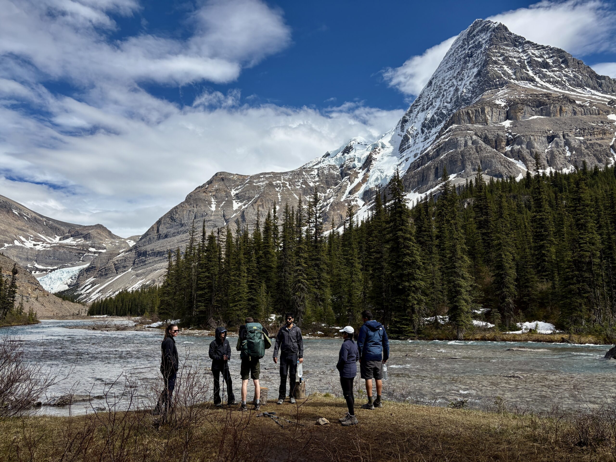Hikers pause beside the Robson River on the Berg Lake Trail with Mount Robson, glaciers, and snow-covered peaks towering above a dense evergreen forest. The scene captures the dramatic mountain landscapes, glacial rivers, and wilderness experience that make the Berg Lake Trail one of Canada's most iconic backpacking routes.