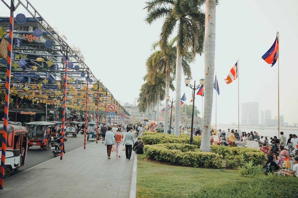 Bustling scene along Phnom Penh riverside with tuk-tuks, people, and flags creating a lively atmosphere.