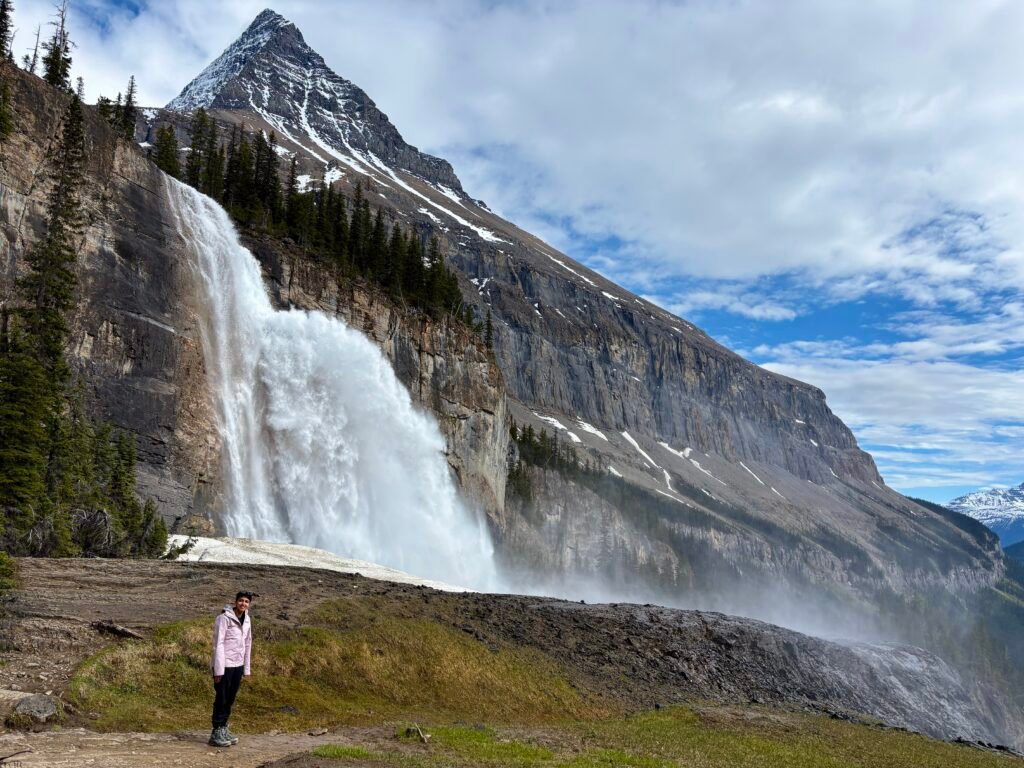 Hiker standing near Emperor Falls on the Berg Lake Trail as a powerful waterfall crashes over a cliff beneath towering snow-streaked mountain peaks. Mist rises from the falls while the dramatic landscape showcases one of the most memorable viewpoints in Mount Robson Provincial Park.