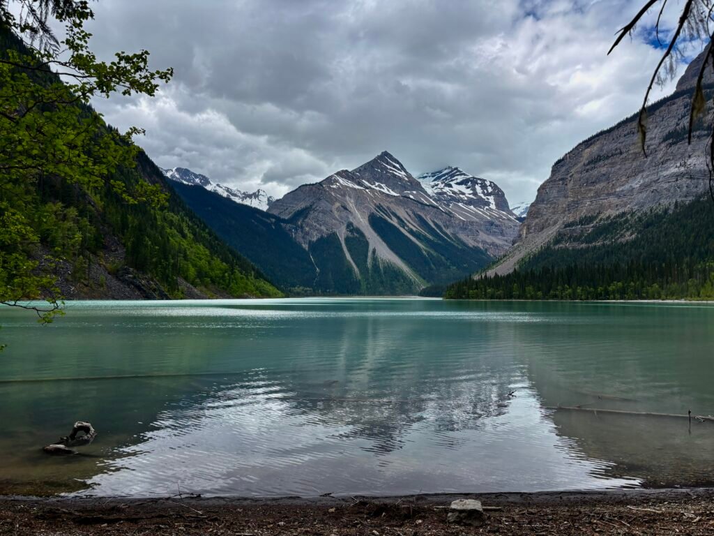Turquoise waters of Kinney Lake reflect snow-capped mountains and forested valley walls along the Berg Lake Trail. Calm water, dramatic Rocky Mountain peaks, and overcast skies create one of the most scenic viewpoints near the start of the hike in Mount Robson Provincial Park.