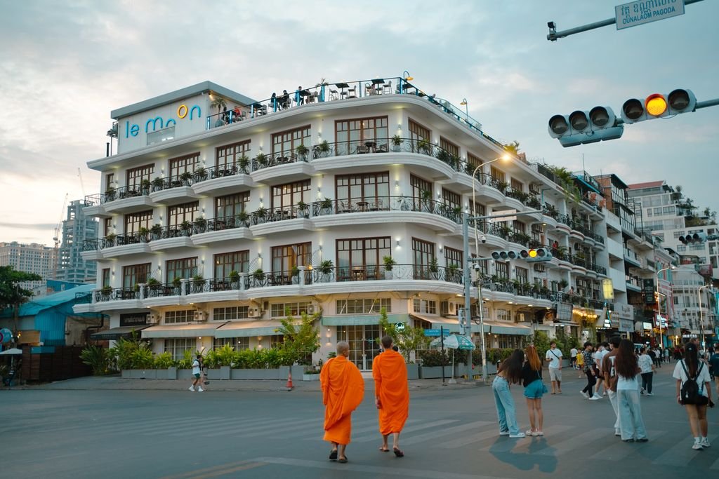 Monks in orange robes walk past a bustling street with modern architecture in Phnom Penh, Cambodia.