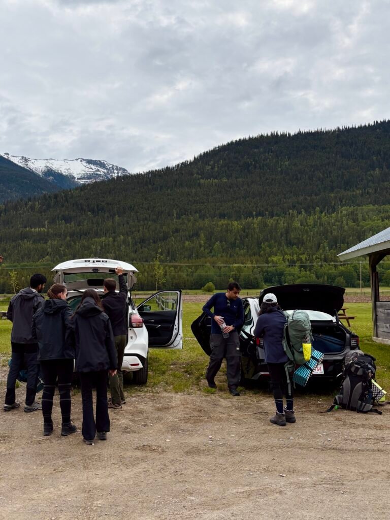 Group of hikers unloading backpacks and organizing camping gear beside two vehicles before beginning the Berg Lake Trail. Forested mountains and snow-capped peaks rise behind them near Mount Robson Provincial Park as they prepare for a multi-day backpacking adventure.
