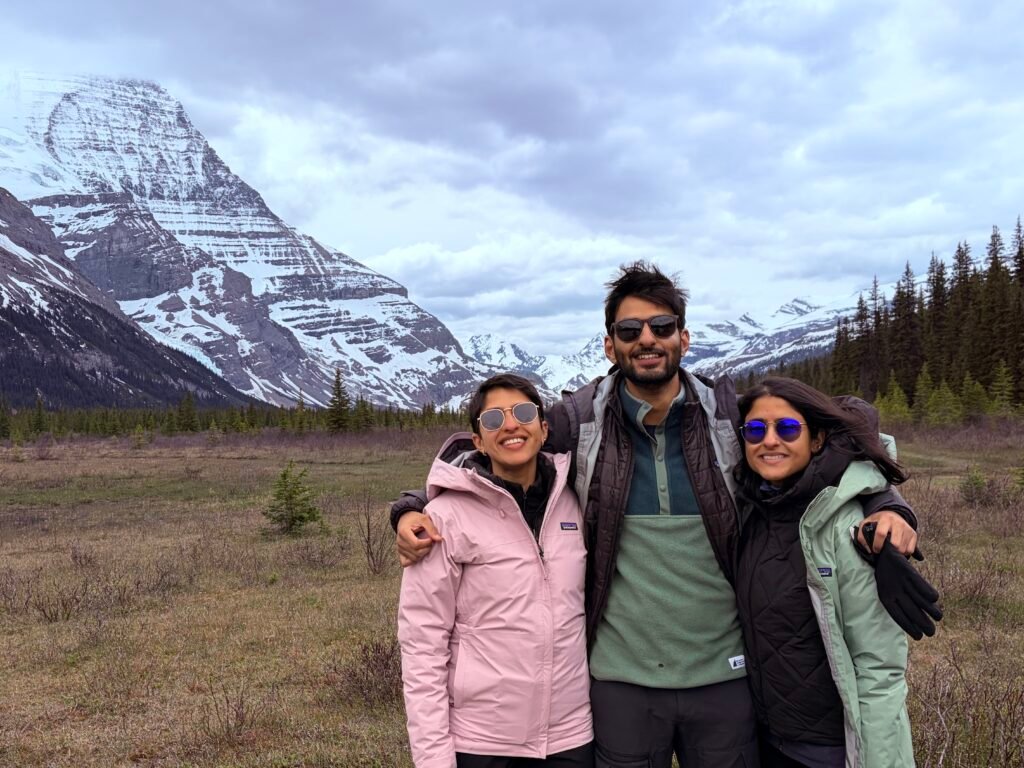 Three hikers stand together in an open alpine meadow near Robson Pass on the Berg Lake Trail, with snow-covered mountains and dramatic Rocky Mountain peaks stretching across the background. The wide valley and glacier-carved landscape showcase the scenery hikers reach at the furthest point of the trail.