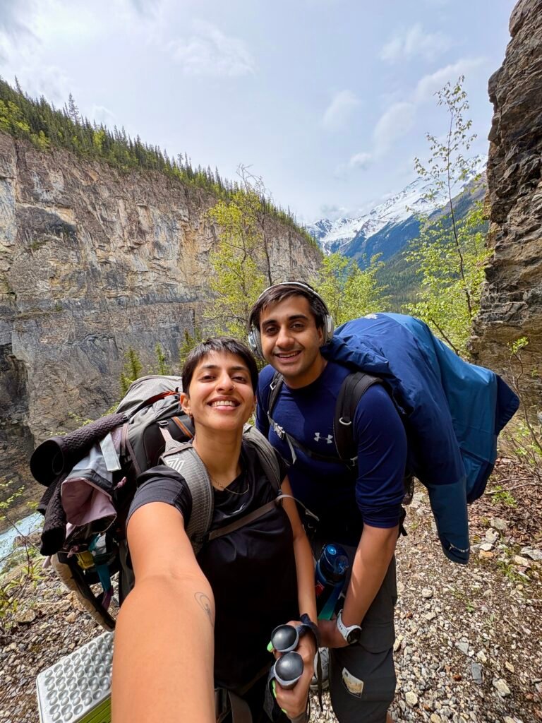 Two backpackers carrying full overnight packs stop for a selfie along the Berg Lake Trail, surrounded by steep canyon walls and snow-covered peaks in the distance. The photo captures the backpacking experience on the trail as hikers make their way through Mount Robson Provincial Park toward Berg Lake.