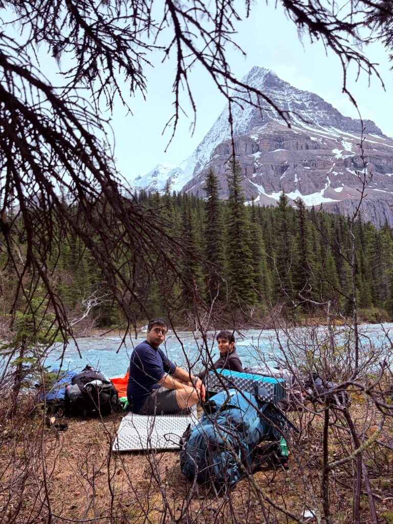 Two backpackers take a break beside a turquoise glacial river on the Berg Lake Trail, surrounded by evergreen forest and a snow-dusted mountain rising in the background. Backpacks and camping gear rest on the ground as they pause to enjoy the scenery during their multi-day hike through Mount Robson Provincial Park.