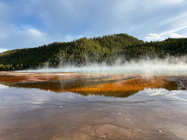 Geothermal spring with steam rising in Yellowstone, reflecting vibrant colors and forests for a 3 day Yellowstone itinerary.