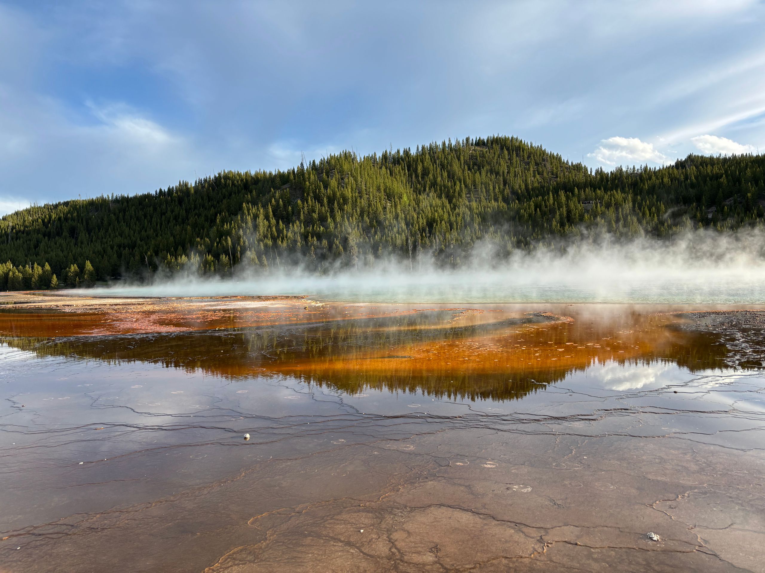 Geothermal spring with steam rising in Yellowstone, reflecting vibrant colors and forests for a 3 day Yellowstone itinerary.