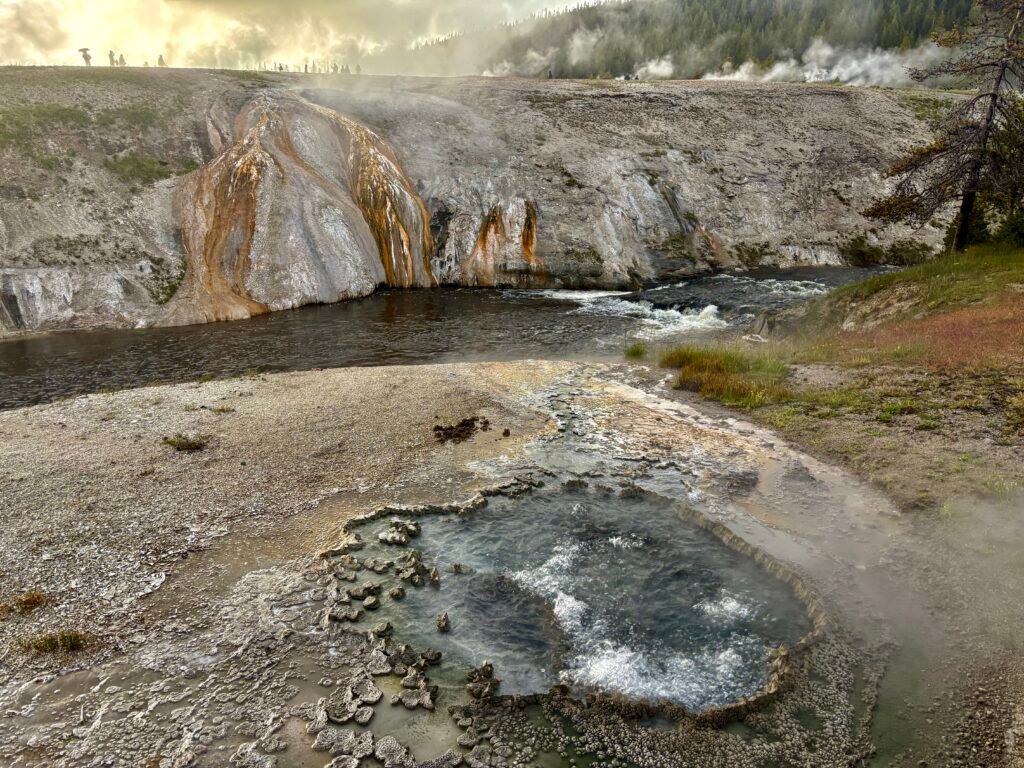 Steaming geothermal pool beside a river in Yellowstone with colorful mineral deposits covering the hillside.