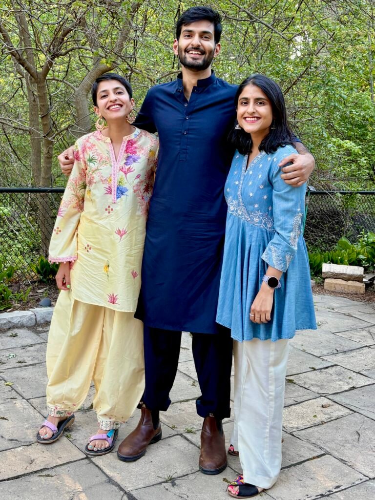 Two women and a man wearing traditional shalwar kameez stand together outdoors with leafy trees in the background. The photo highlights traditional Pakistani clothing in a natural setting.